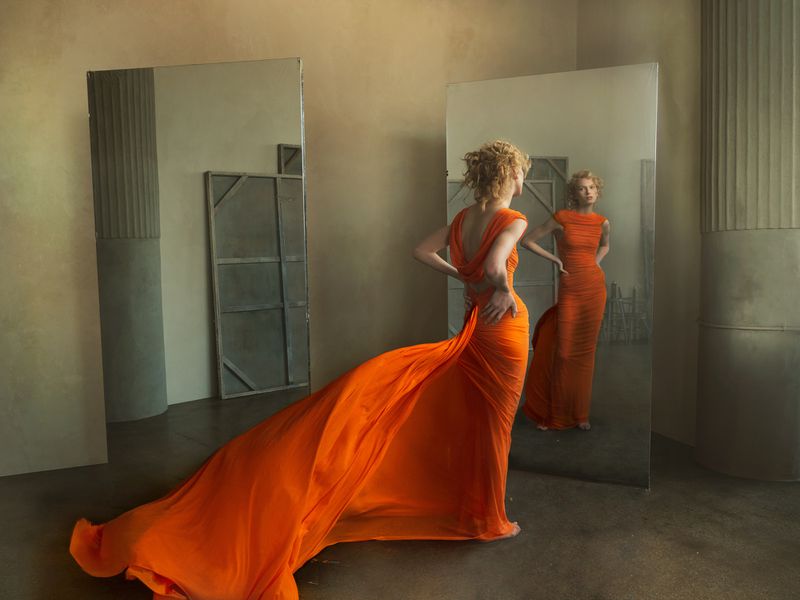 Woman in orange gown poses near mirror, reflecting her image. Soft light illuminates the studio space with architectural details.