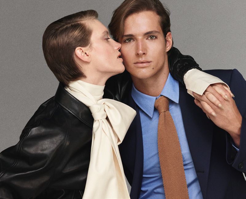A fashionable couple poses indoors; the woman is whispering into the man's ear, both wearing formal attire in a studio setting.