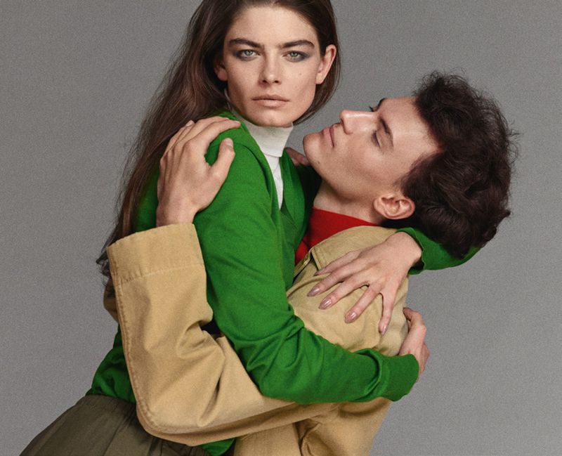 Fashion shot of a stylish Caucasian couple in a close embrace. The woman has long brown hair and wears a green sweater and turtleneck.