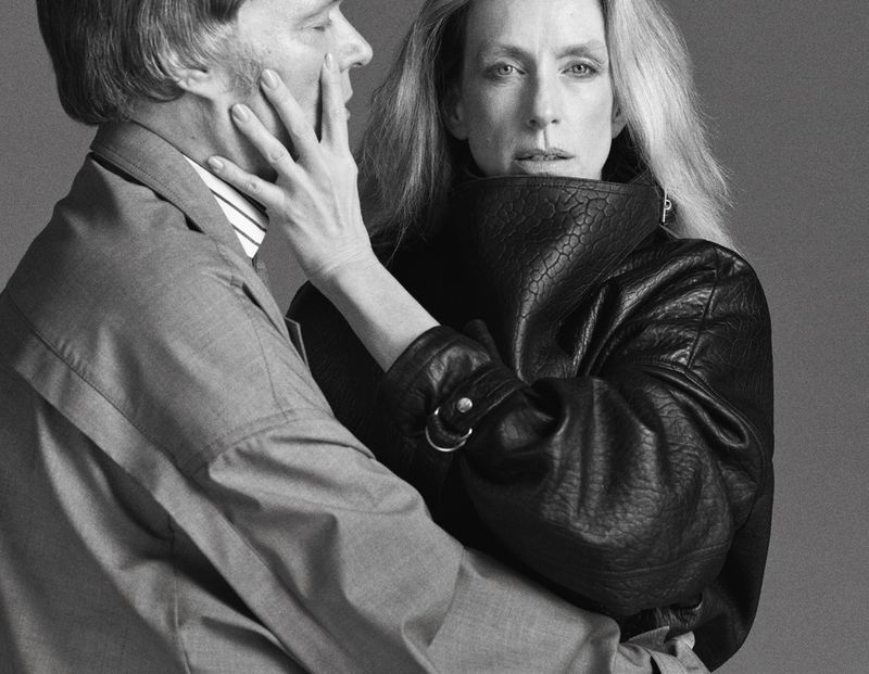 Monochrome portrait of a woman in a textured leather jacket and a man in a suit with a hand on his face, against a gray background.
