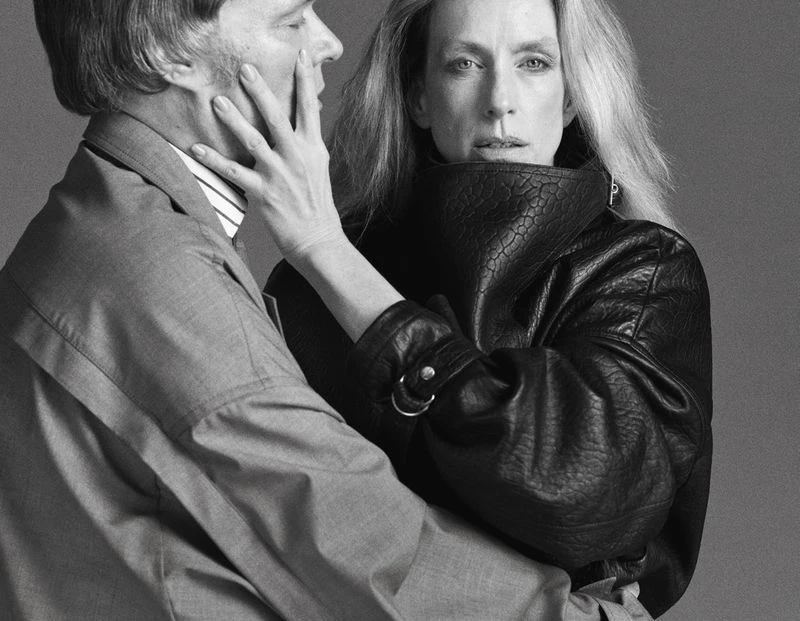 Monochrome portrait of a woman in a textured leather jacket and a man in a suit with a hand on his face, against a gray background.