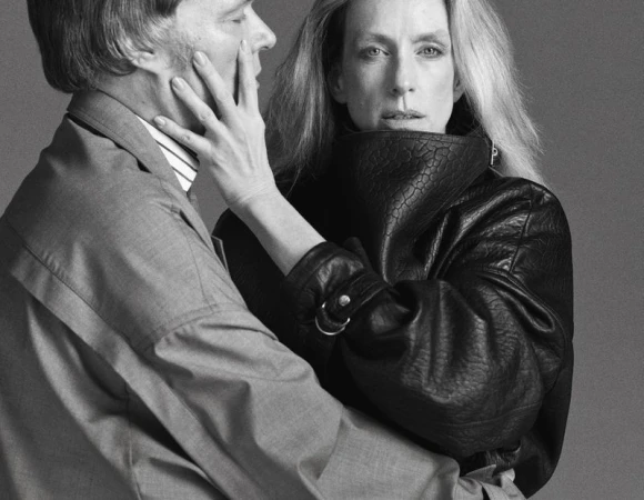 Monochrome portrait of a woman in a textured leather jacket and a man in a suit with a hand on his face, against a gray background.