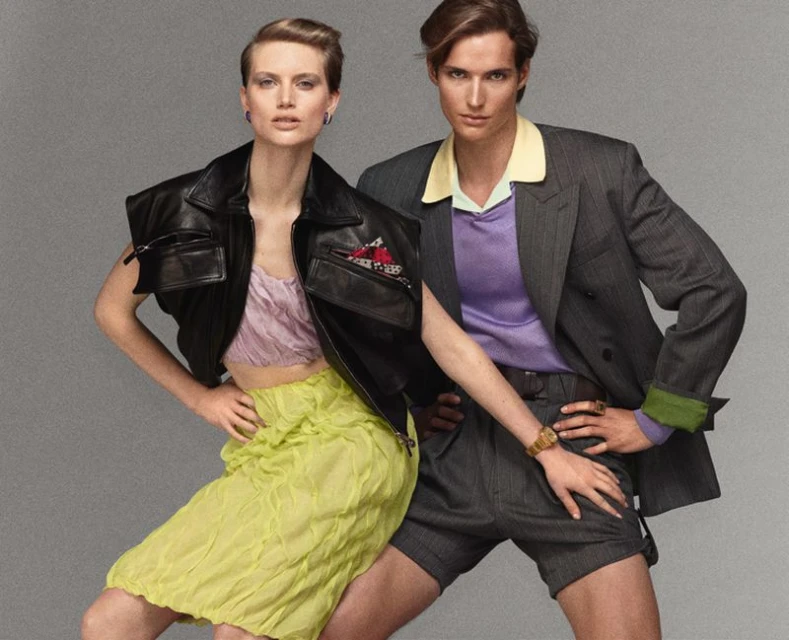 Two fashion models in a photo studio. The woman wears a black vest and green skirt while the man is in a pinstripe suit and shorts.