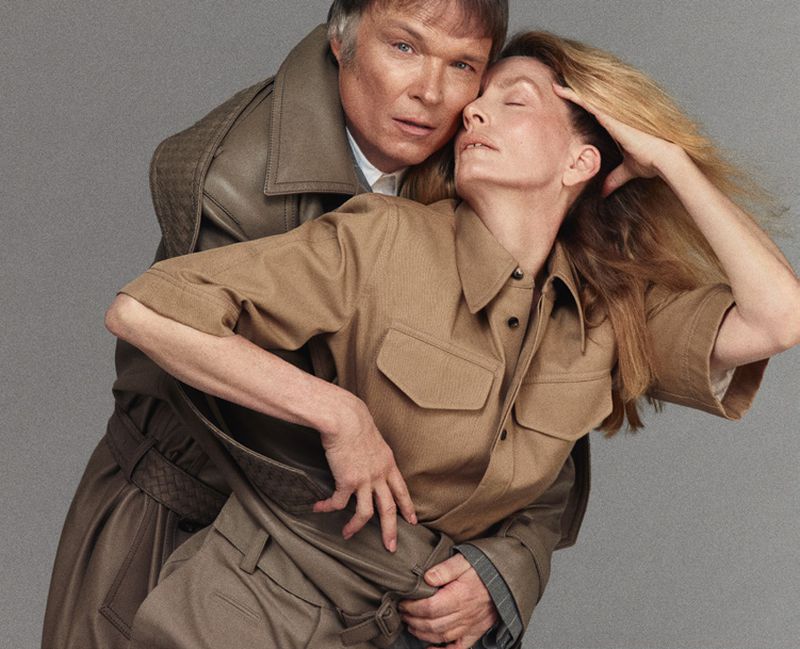 Adult male and female embrace indoors, wearing fashionable clothing with neutral color palette in a studio setting with plain gray backdrop.