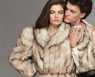 Woman and man posing closely in plush beige fur coats against a neutral grey studio backdrop.