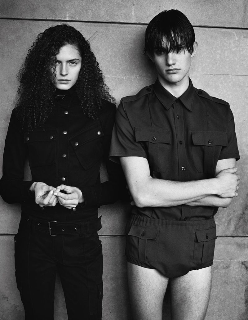 Monochrome studio shot of a female model with curly hair and a male model in a button-down shirt with short bottoms posing indoors.