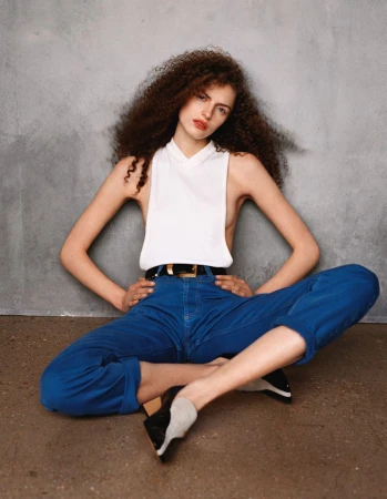 A young woman with long curly hair in a white sleeveless top and blue jeans sits on the floor against a textured gray wall.