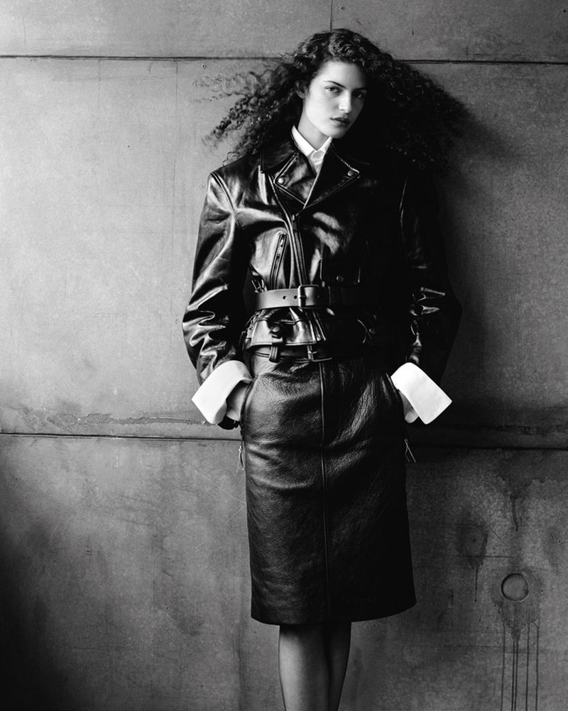 Model with long dark curly hair wearing a leather jacket and skirt against a concrete wall