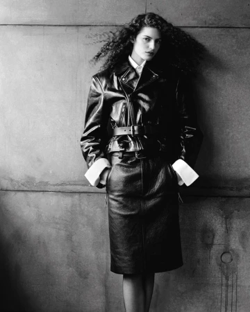 Model with long dark curly hair wearing a leather jacket and skirt against a concrete wall