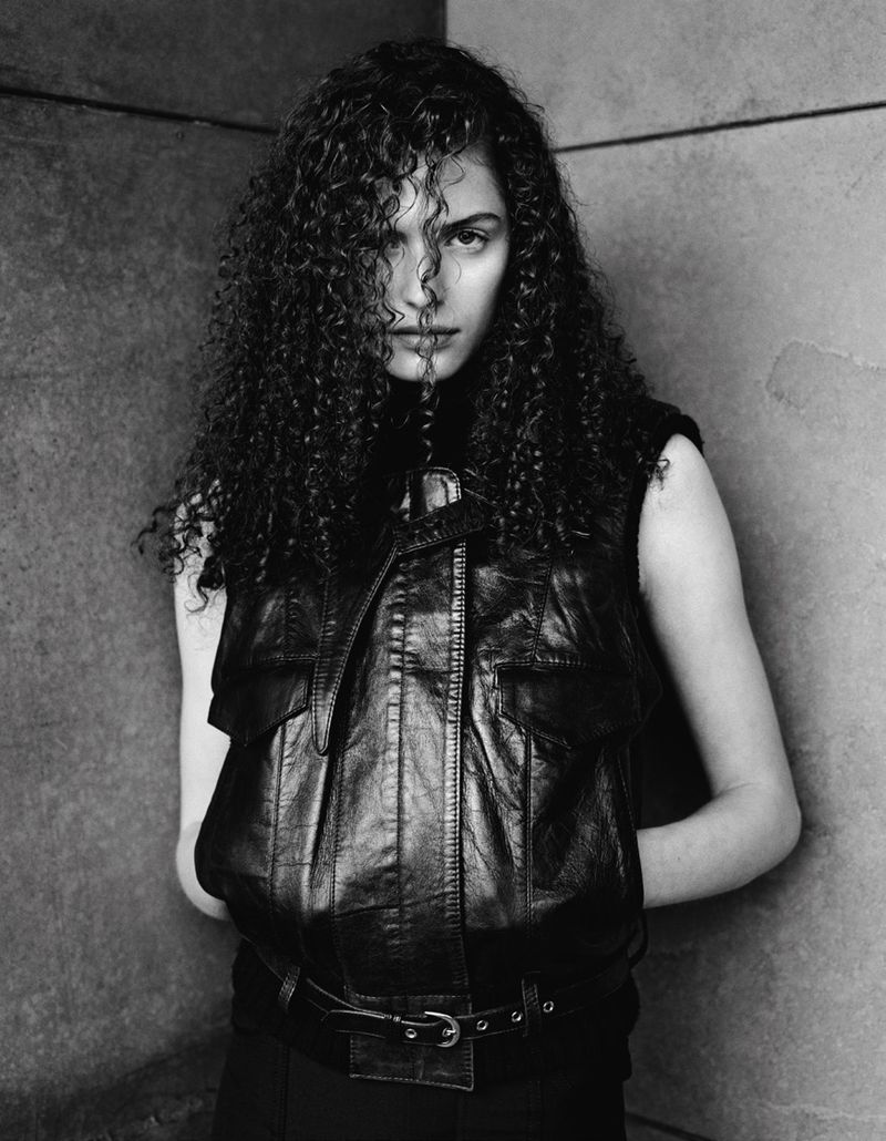 Monochrome portrait of a woman with curly hair, wearing a black leather vest, against a concrete wall backdrop, styled in editorial fashion.
