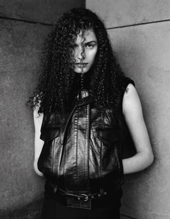 Monochrome portrait of a woman with curly hair, wearing a black leather vest, against a concrete wall backdrop, styled in editorial fashion.