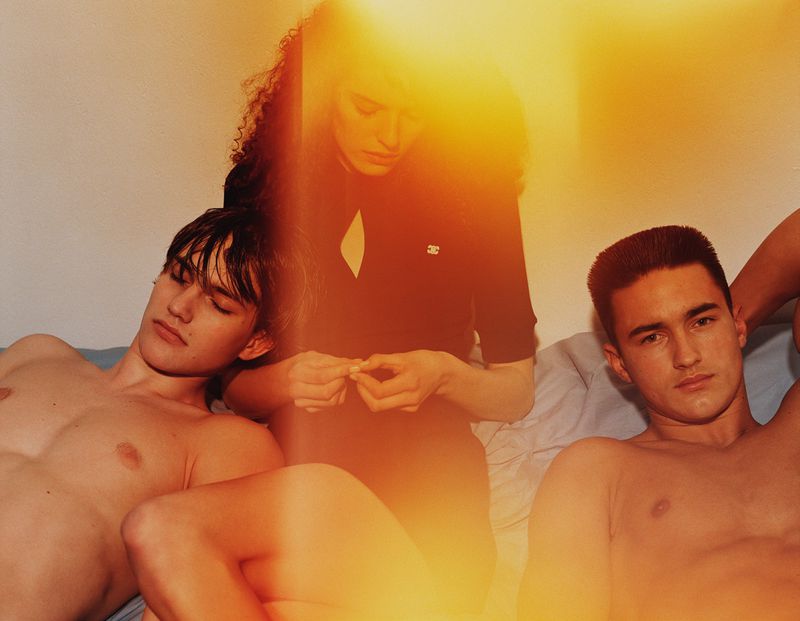 Three people pose indoors on a bed with a bright light flare obscuring some details, one woman and two men.