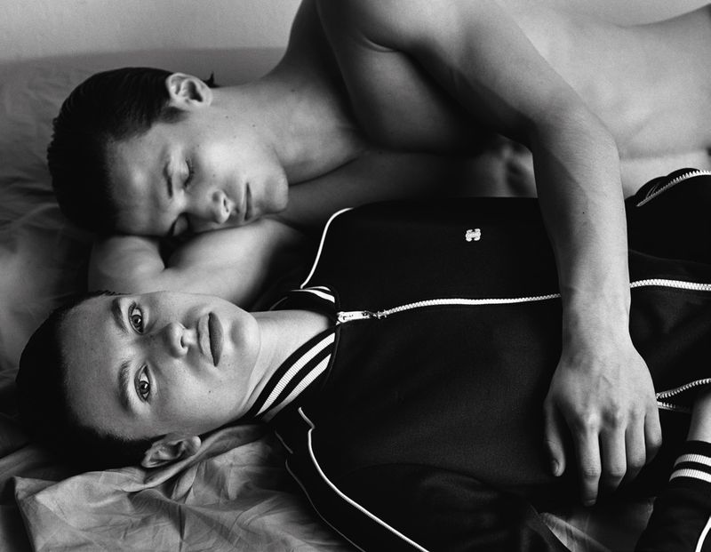 Black and white portrait of two young adult males lying on a bed; one is sleeping, while the other is looking directly at the camera.
