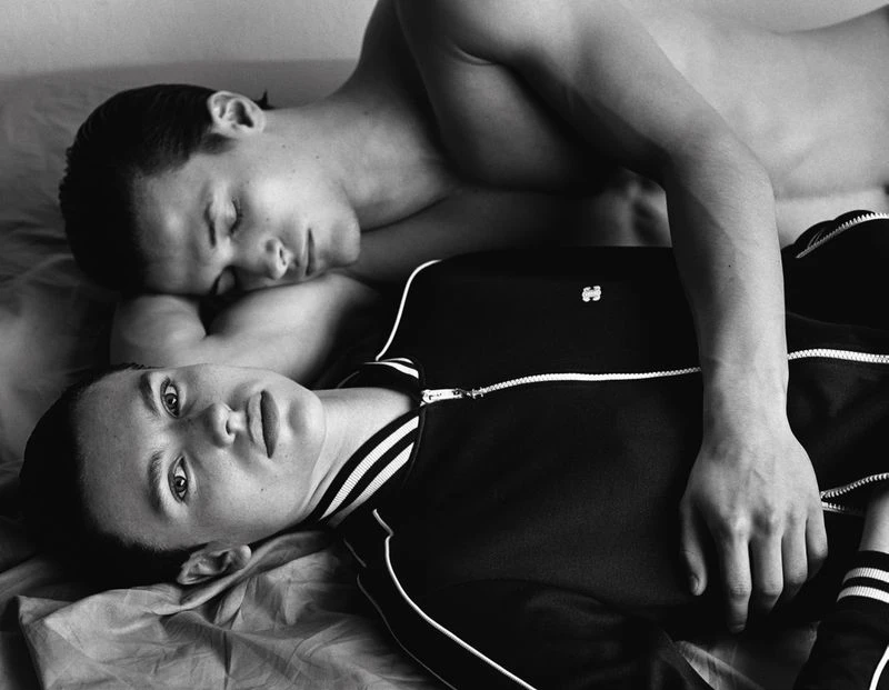 Black and white portrait of two young adult males lying on a bed; one is sleeping, while the other is looking directly at the camera.