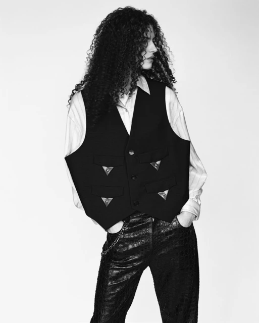 Monochrome studio shot of a young adult with curly hair, wearing a vest with metal accents and textured pants against a white backdrop.