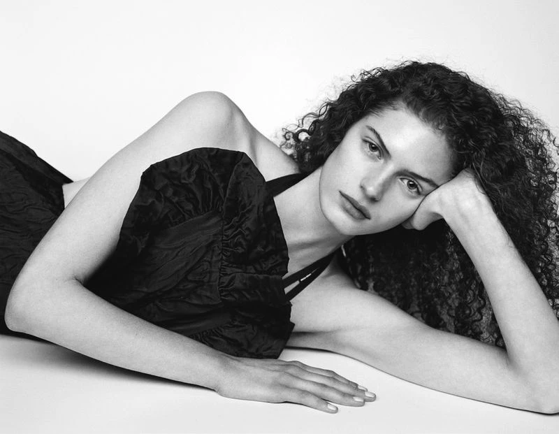 A young woman with curly hair is lying down in a black dress. She is indoors with a white background and is posing for a portrait.