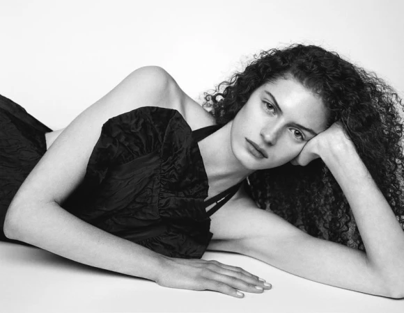 A young woman with curly hair is lying down in a black dress. She is indoors with a white background and is posing for a portrait.