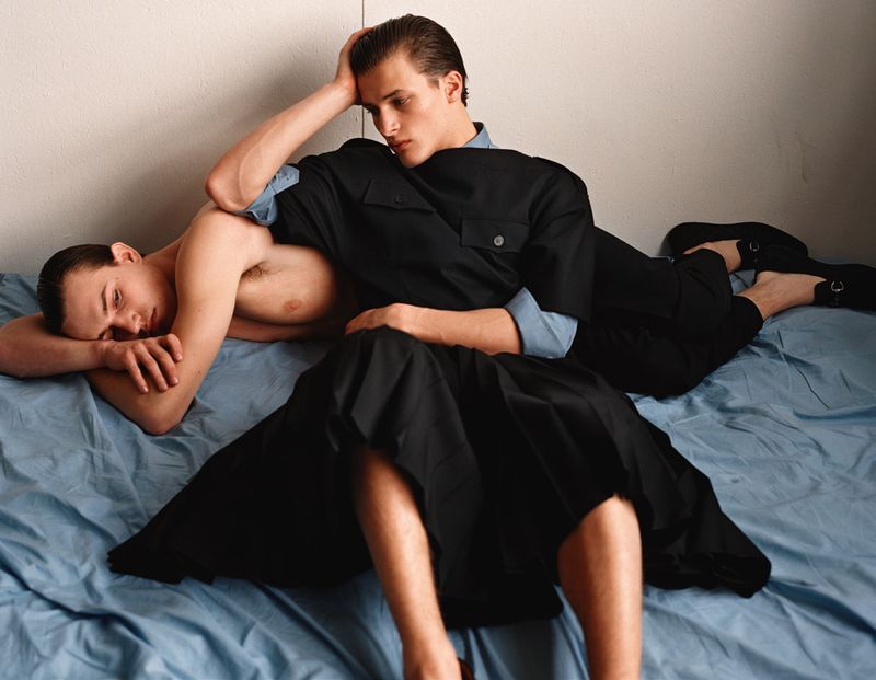 Two young men lying on a blue bed sheet, one shirtless, the other in a black outfit, indoors with neutral wall for fashion editorial.
