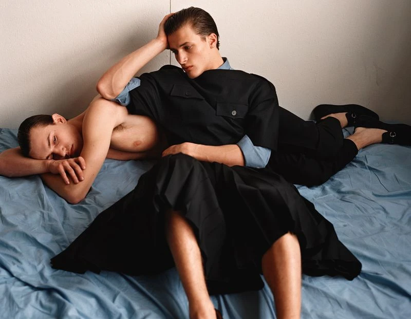 Two young men lying on a blue bed sheet, one shirtless, the other in a black outfit, indoors with neutral wall for fashion editorial.