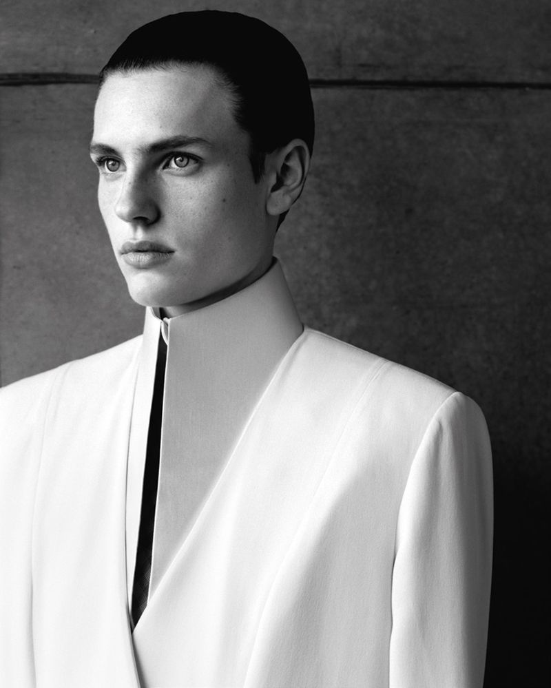 Monochrome portrait of a model with slicked-back hair wearing a structured high-collar white jacket.