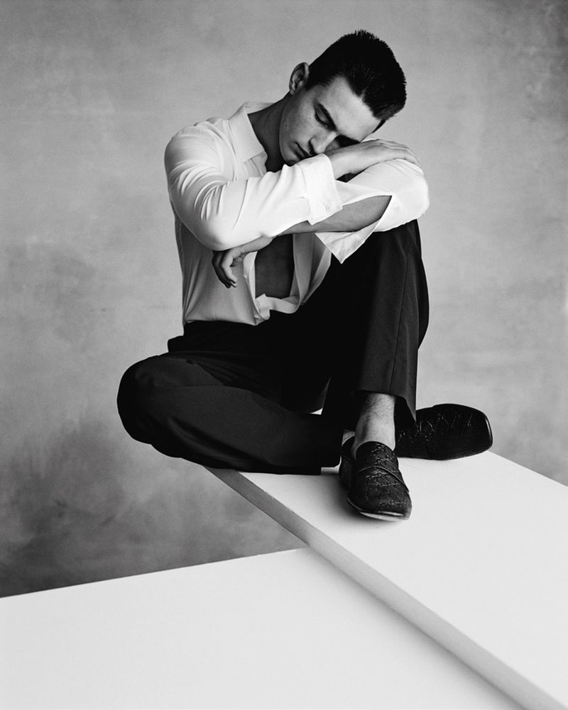Monochrome studio portrait of a dark-haired adult man in a white dress shirt and dark trousers, sitting in a pensive pose on a white platform.