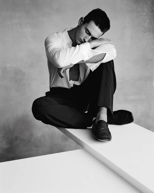 Monochrome studio portrait of a dark-haired adult man in a white dress shirt and dark trousers, sitting in a pensive pose on a white platform.