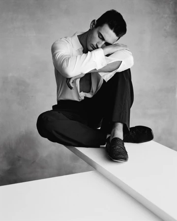 Monochrome studio portrait of a dark-haired adult man in a white dress shirt and dark trousers, sitting in a pensive pose on a white platform.
