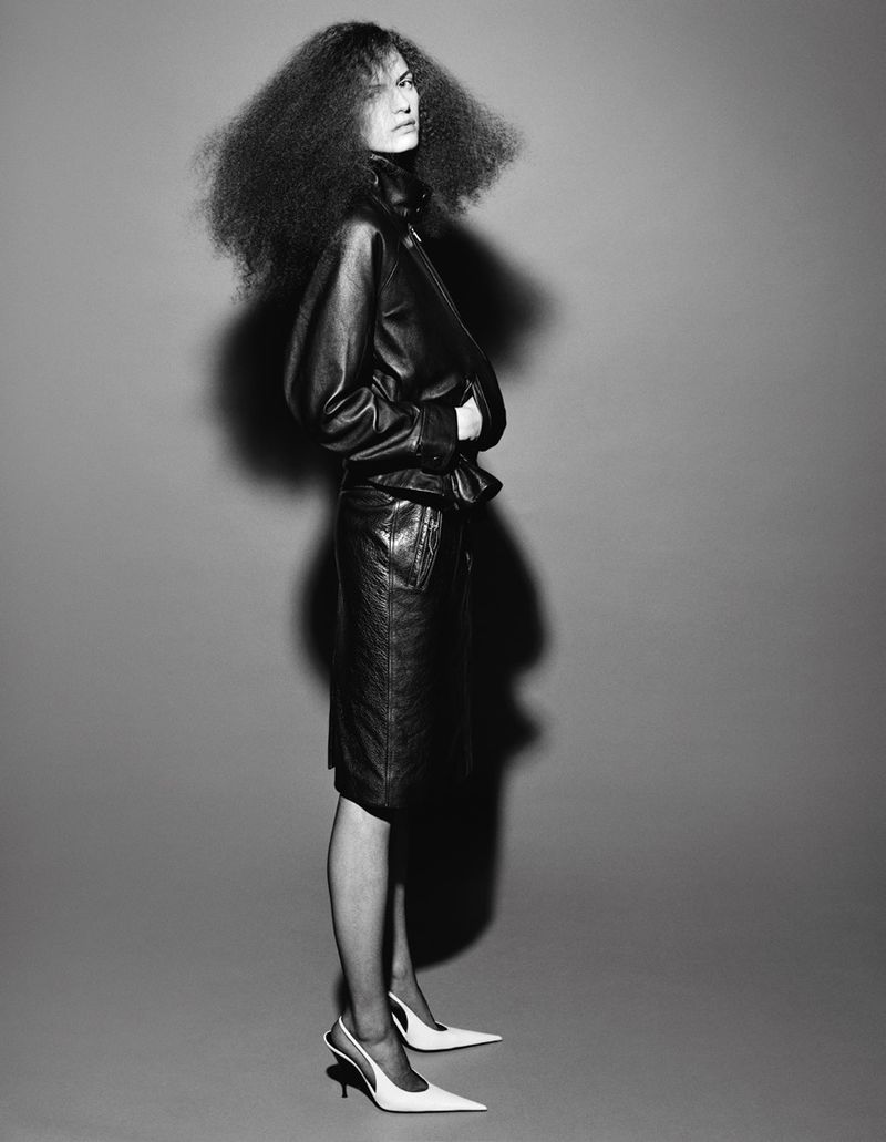 Model with voluminous textured hair wearing a black leather jacket and skirt against a grey background