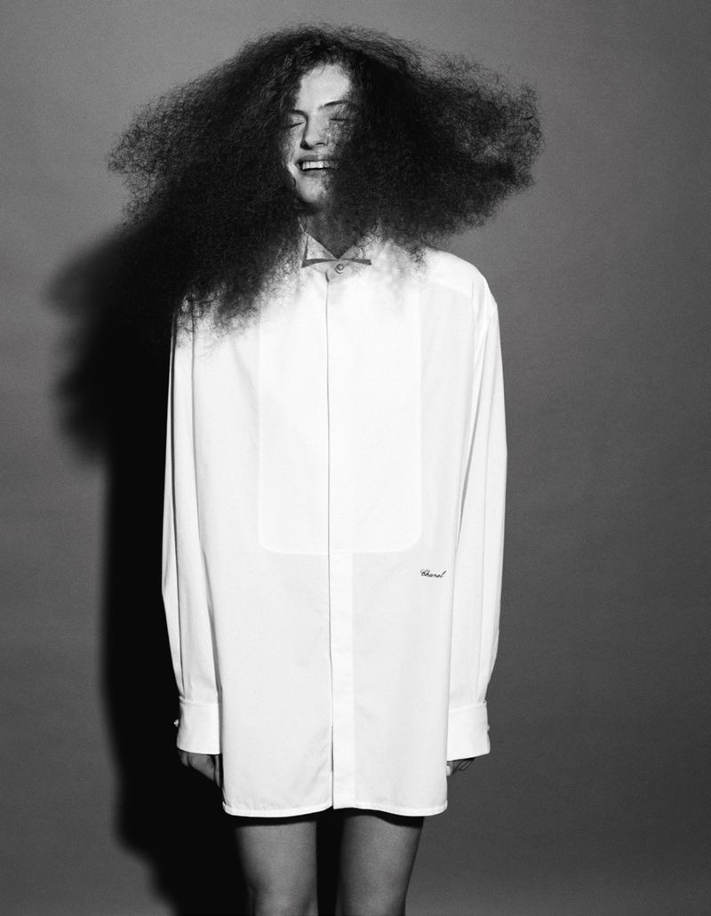 Portrait of a happy adult wearing a large, white dress shirt, standing in front of a grey studio backdrop with very large, curly dark hair.