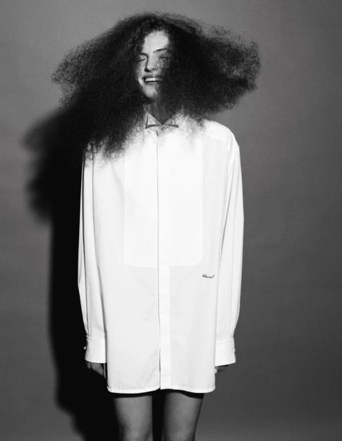 Portrait of a happy adult wearing a large, white dress shirt, standing in front of a grey studio backdrop with very large, curly dark hair.