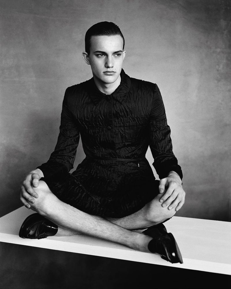 Monochrome studio portrait of a slim male fashion model sitting cross-legged on a white platform wearing a dark textured jacket and loafers.