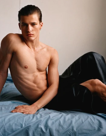 Male model posing shirtless in black leather pants on a blue sheet. Indoor studio shot with neutral background, demonstrating fashion and style.