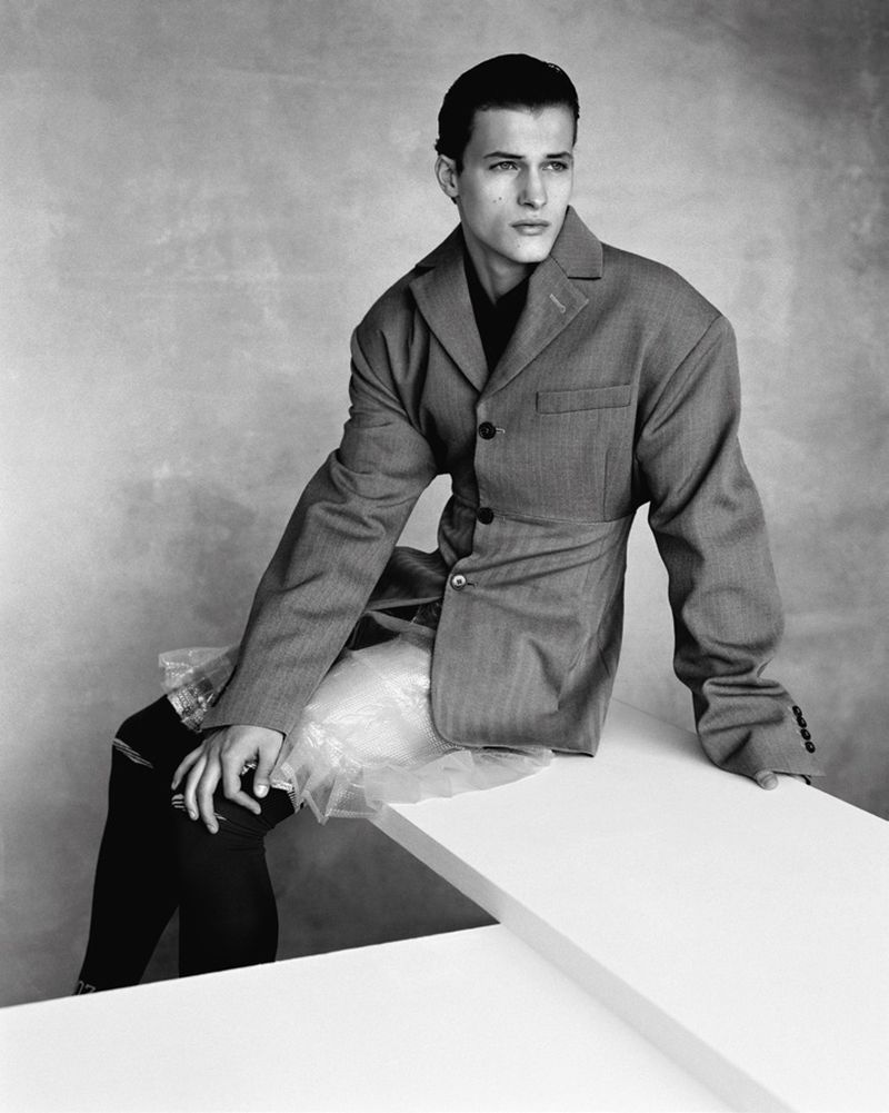 Monochrome portrait of a male model in a blazer and skirt, seated against a textured backdrop with a contemplative gaze.