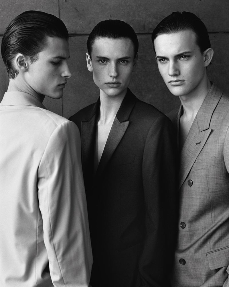 Three men in suits stand closely together against a grey background in a monochromatic fashion shot, looking stylish and well-groomed.