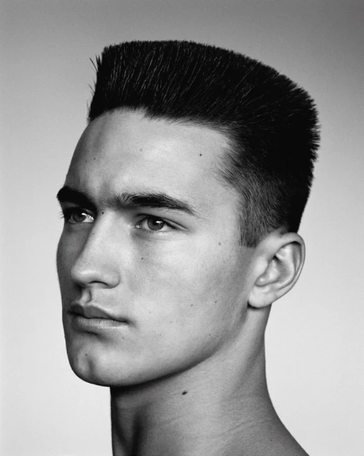 Black and white portrait of a young adult male with a flattop haircut, gazing off-camera against a neutral background in a photo studio.