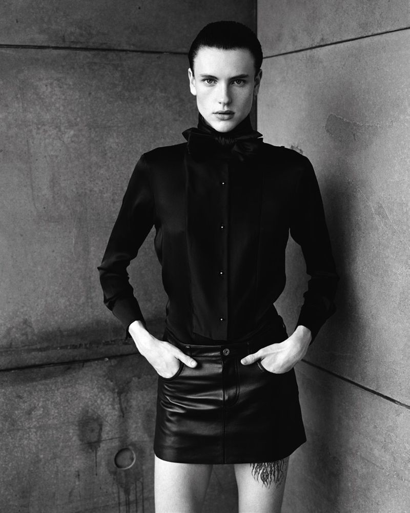 Studio shot of an adult with short dark hair, posing in black button-down shirt and a black leather skirt against a grey concrete wall.
