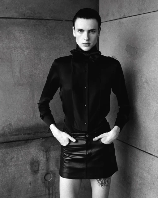 Studio shot of an adult with short dark hair, posing in black button-down shirt and a black leather skirt against a grey concrete wall.