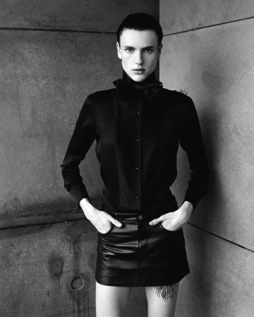 Studio shot of an adult with short dark hair, posing in black button-down shirt and a black leather skirt against a grey concrete wall.