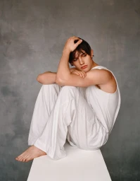 Young man seated barefoot on a white block wearing a white tank top and loose trousers against a textured grey studio backdrop.