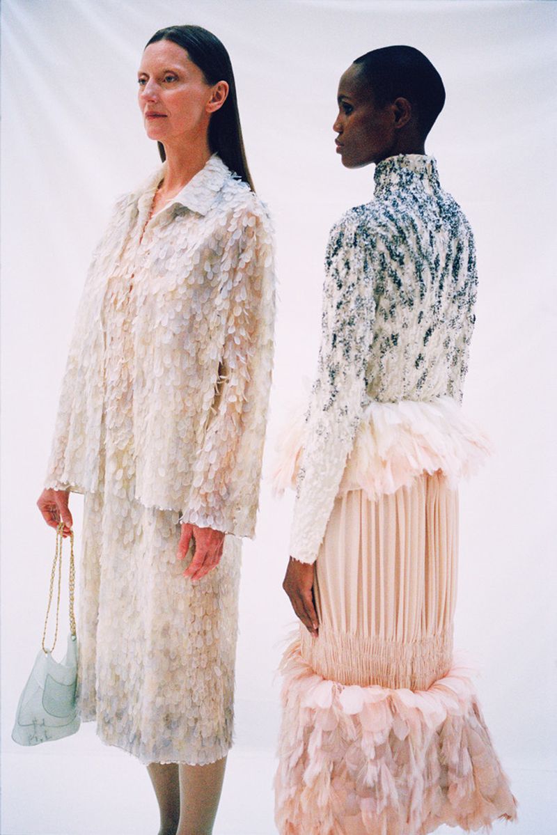 Two women stand side-by-side in an indoor space, wearing couture gowns in neutral tones of white and peach, accented with floral and beaded details.