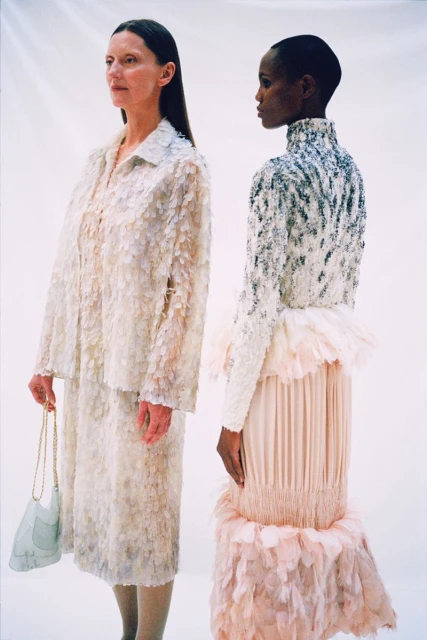 Two women stand side-by-side in an indoor space, wearing couture gowns in neutral tones of white and peach, accented with floral and beaded details.
