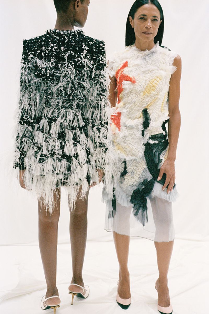 Two women model avant-garde fashion dresses in a bright studio. The dresses feature tiered, feathery textures and bold patterns, accessorized with pumps.