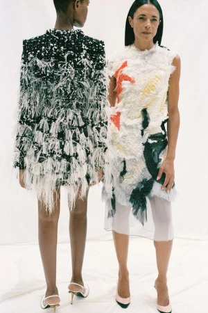 Two women model avant-garde fashion dresses in a bright studio. The dresses feature tiered, feathery textures and bold patterns, accessorized with pumps.