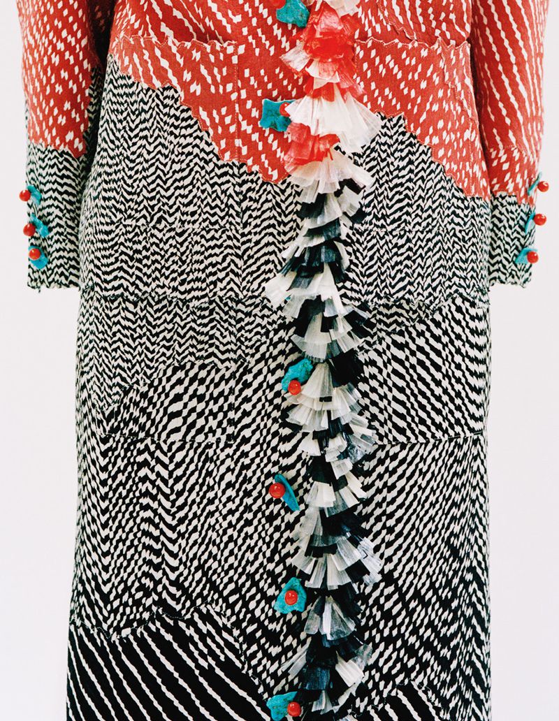 Close-up of red and black and white patterned dress with ruffled trim and decorative turquoise and orange accents against white.