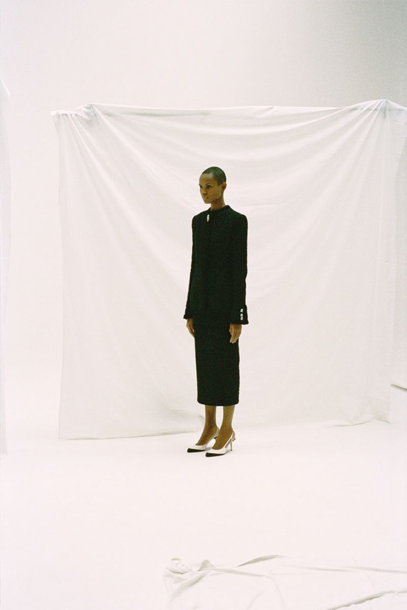 A woman with short hair in a black skirt suit and cream-colored heels stands in a white photo studio with soft lighting.