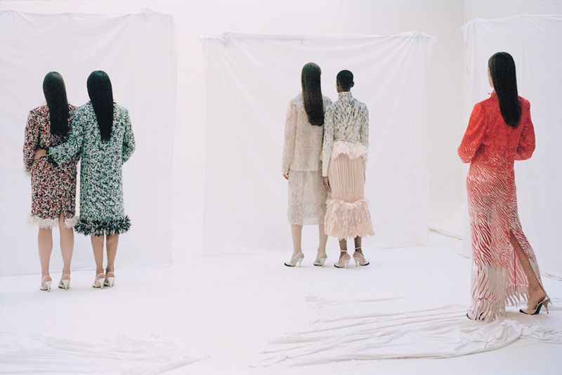 Five models posing with their backs turned in textured designer garments against draped white fabric backdrops in a photo studio.