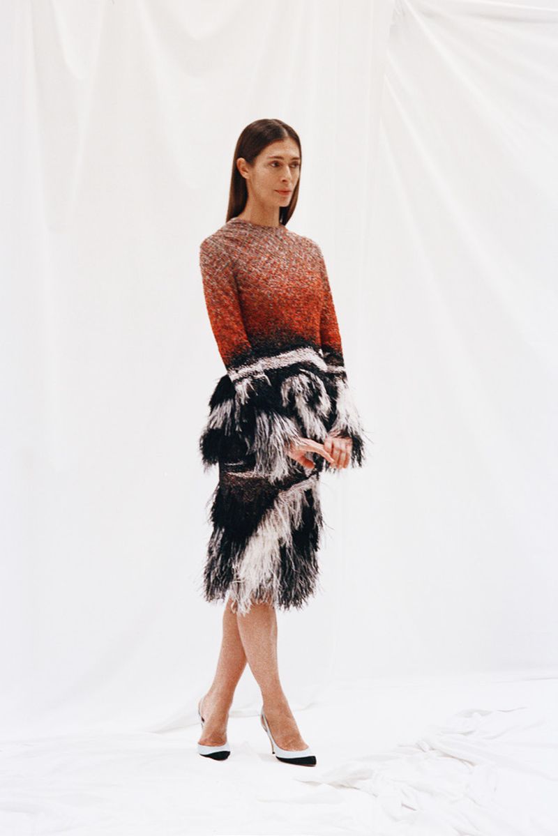 A woman poses in a long-sleeved, ombre dress with black and white fringe. She is wearing heels and is standing against a white backdrop.