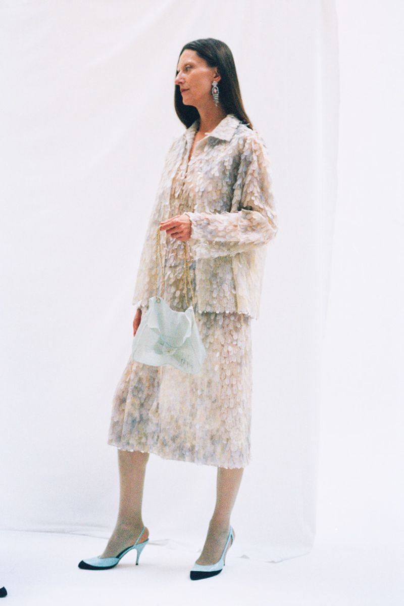 An adult female model poses in a studio wearing a floral-patterned skirt and jacket, light-colored hosiery, slingback heels, and dangling earrings.