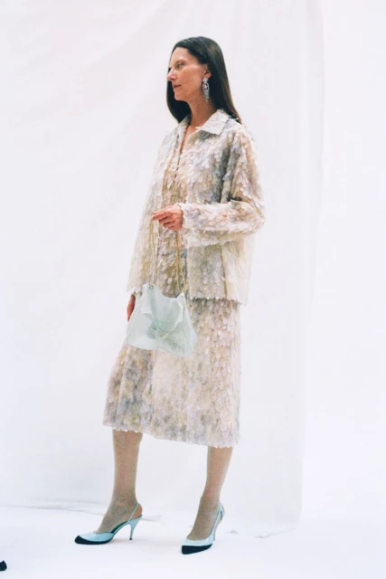 An adult female model poses in a studio wearing a floral-patterned skirt and jacket, light-colored hosiery, slingback heels, and dangling earrings.