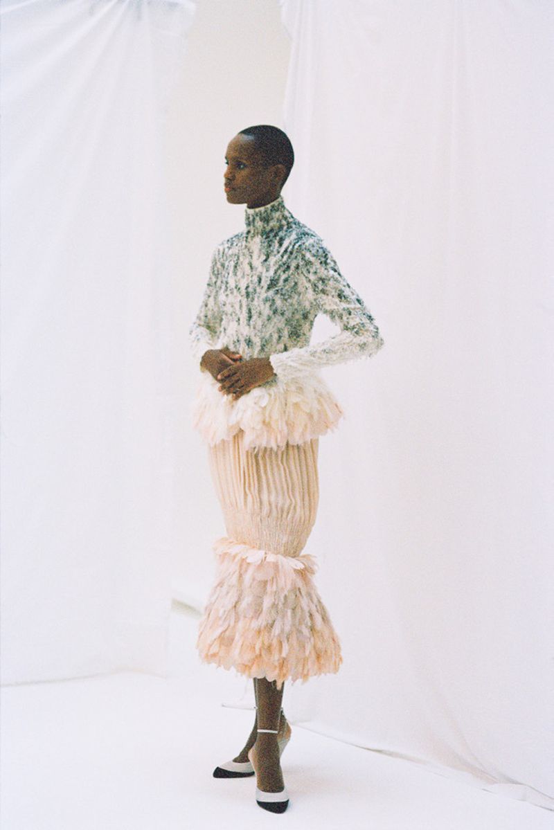 A Black female fashion model poses in a white studio wearing a unique feathered dress with a long-sleeved embellished turtleneck top.
